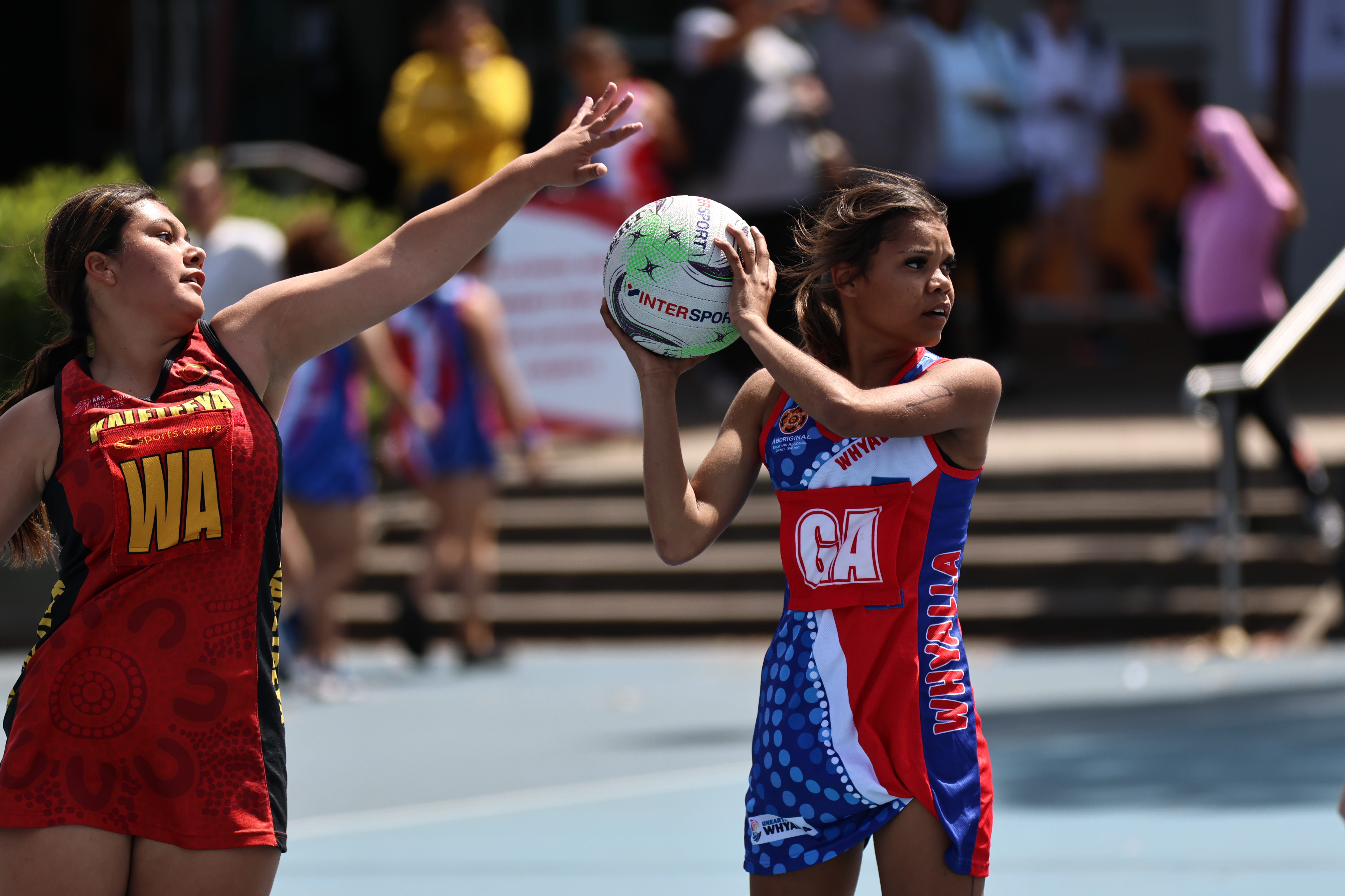 Player holding the ball on her shoulder looking to pass it whilst being defended by opposition player with her arm out. 